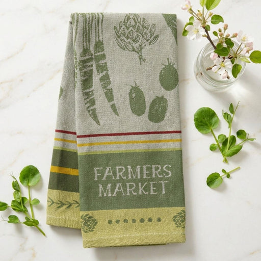 Kitchen towel with 'Farmers Market' text and vegetable patterns on a marble surface with greenery.