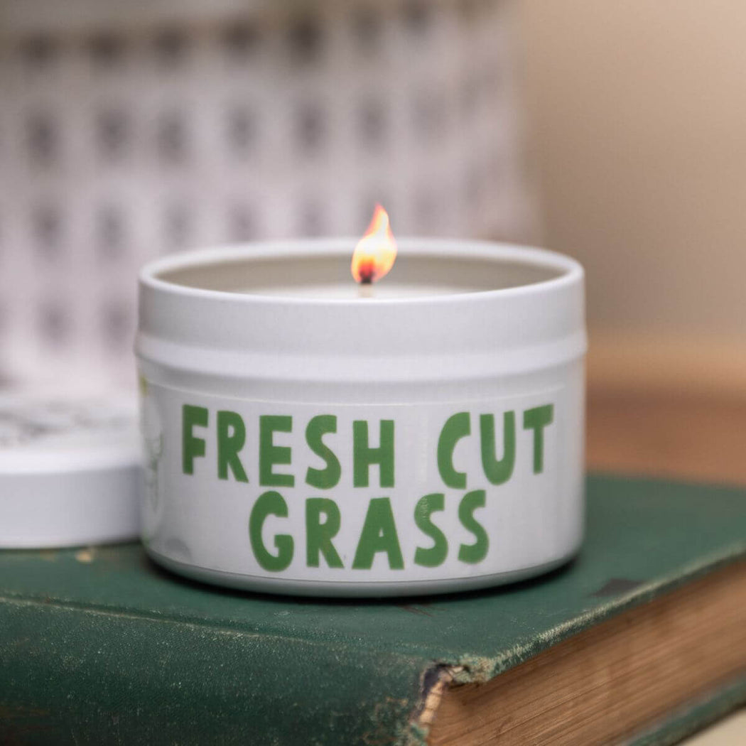 Candle in a container labeled 'Fresh Cut Grass' on a surface with a blurred background