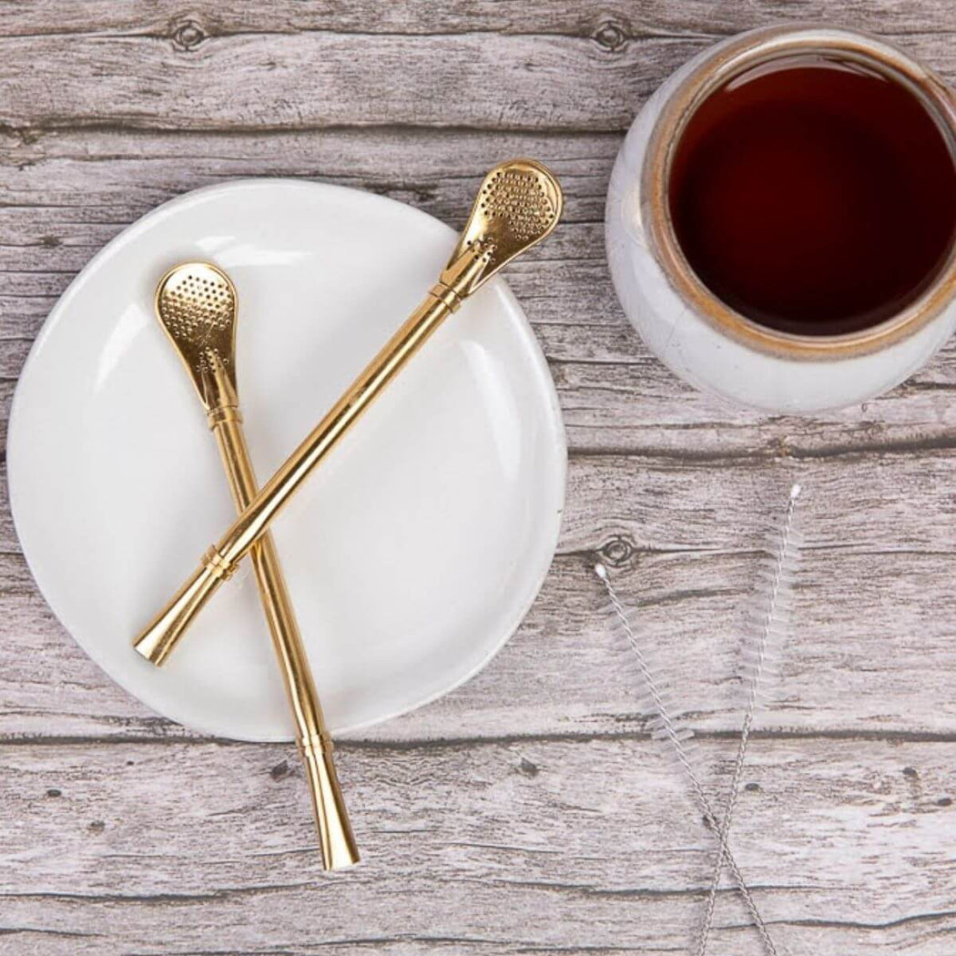 Two gold filter straws on a white plate next to a cup of tea on a wooden surface.