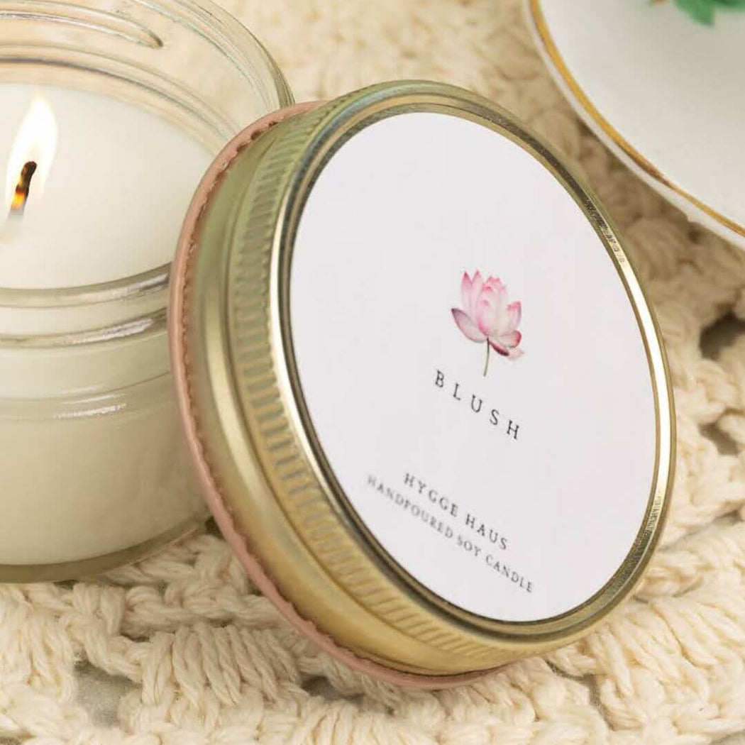 Candle with a 'Blush' lid featuring a pink flower design on a textured surface.