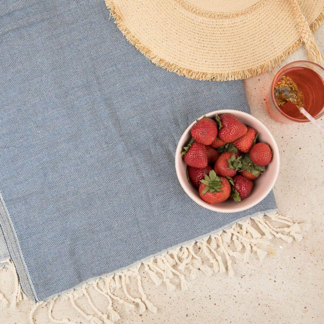 A styled flat-lay of a light blue woven textile with cream fringe, arranged on a light beige surface alongside a bowl of strawberries, a cup of iced tea, and part of a straw hat.