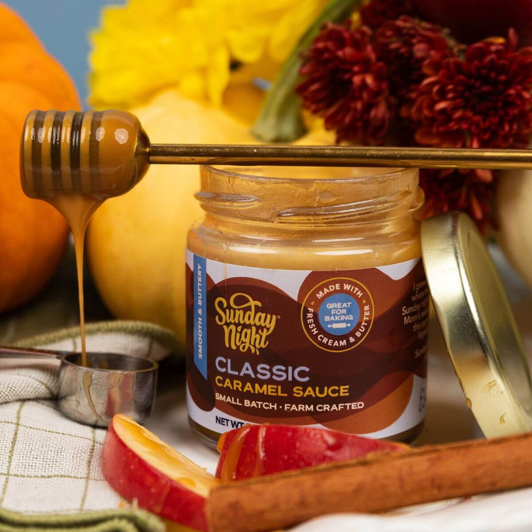 A jar of Sunday Night Classic Caramel Sauce with the lid off, a honey dipper resting across the jar, and autumn ingredients like apples, cinnamon, a metal measuring cup, and flowers in the background.