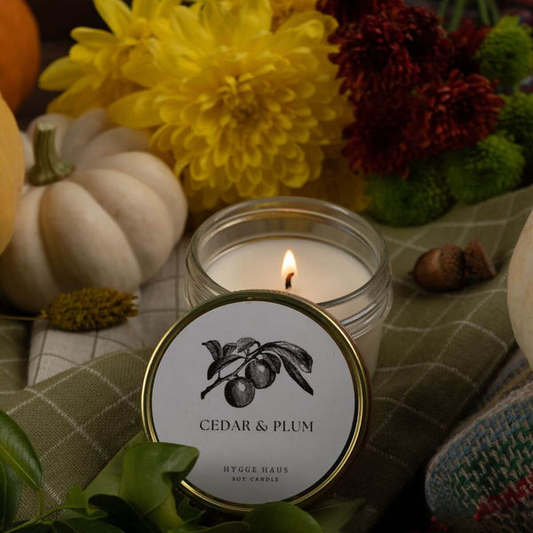 A lit scented candle in a clear glass jar sits on a plaid fabric surface with a round lid in front showing the label 'CEDAR & PLUM' and 'HYGGE HAUS SCENT CANDLE.' The scene is styled with pumpkins, flowers, leaves, and acorns in an autumn setting.