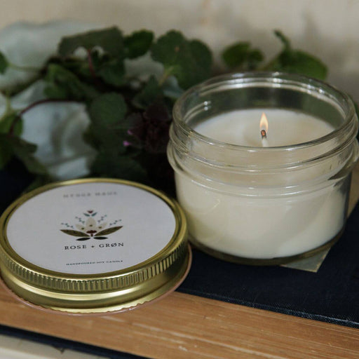 Candle in a glass jar with a lit wick, placed on a wooden surface with a blurred natural background.