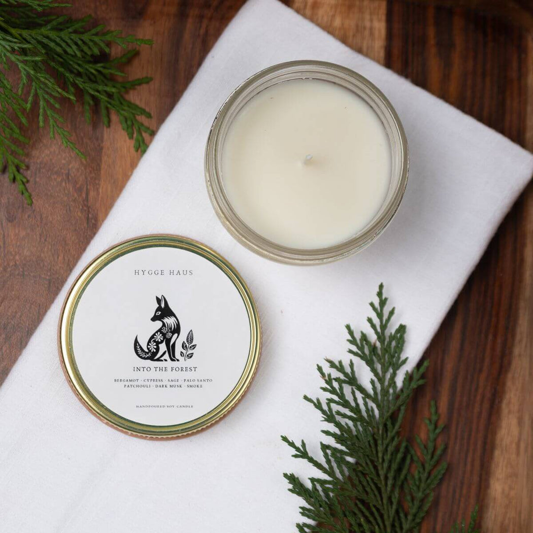 Candle in a glass jar with a lid featuring a label with the words "Into the Forest" laid on a white cloth with greenery around.