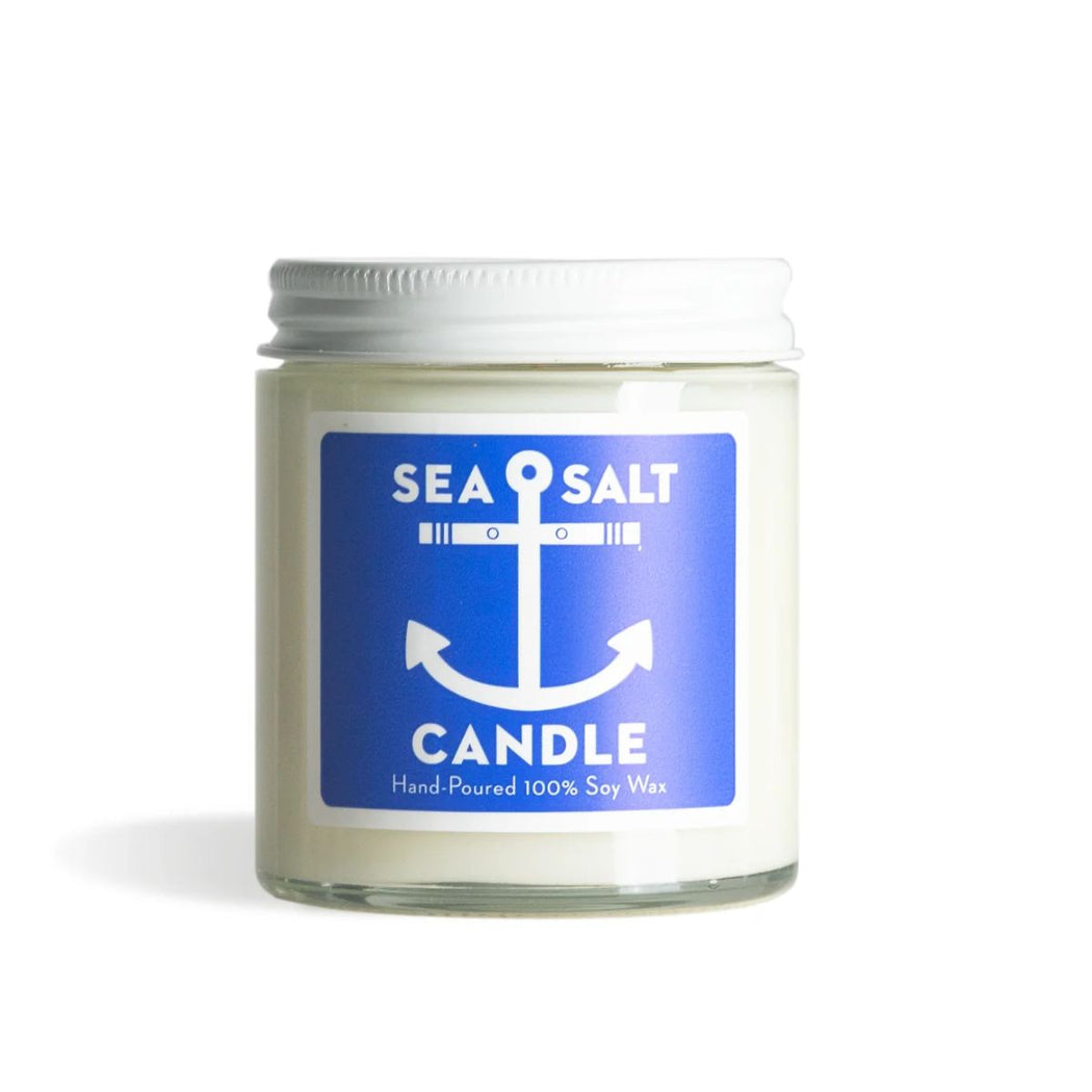 A sea salt scented soy wax candle in a clear glass jar with a white metal lid and a blue label featuring a white anchor graphic.