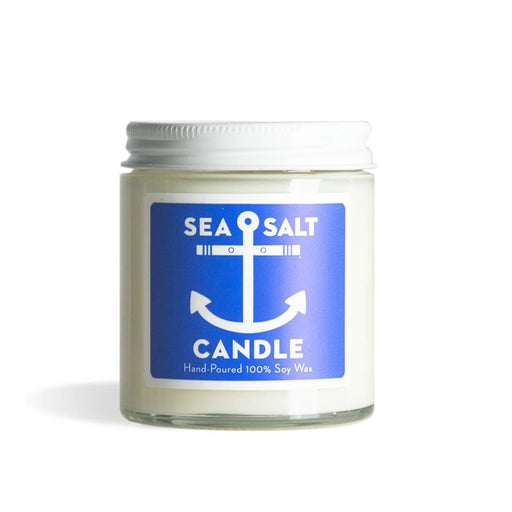 A sea salt scented soy wax candle in a clear glass jar with a white metal lid and a blue label featuring a white anchor graphic.