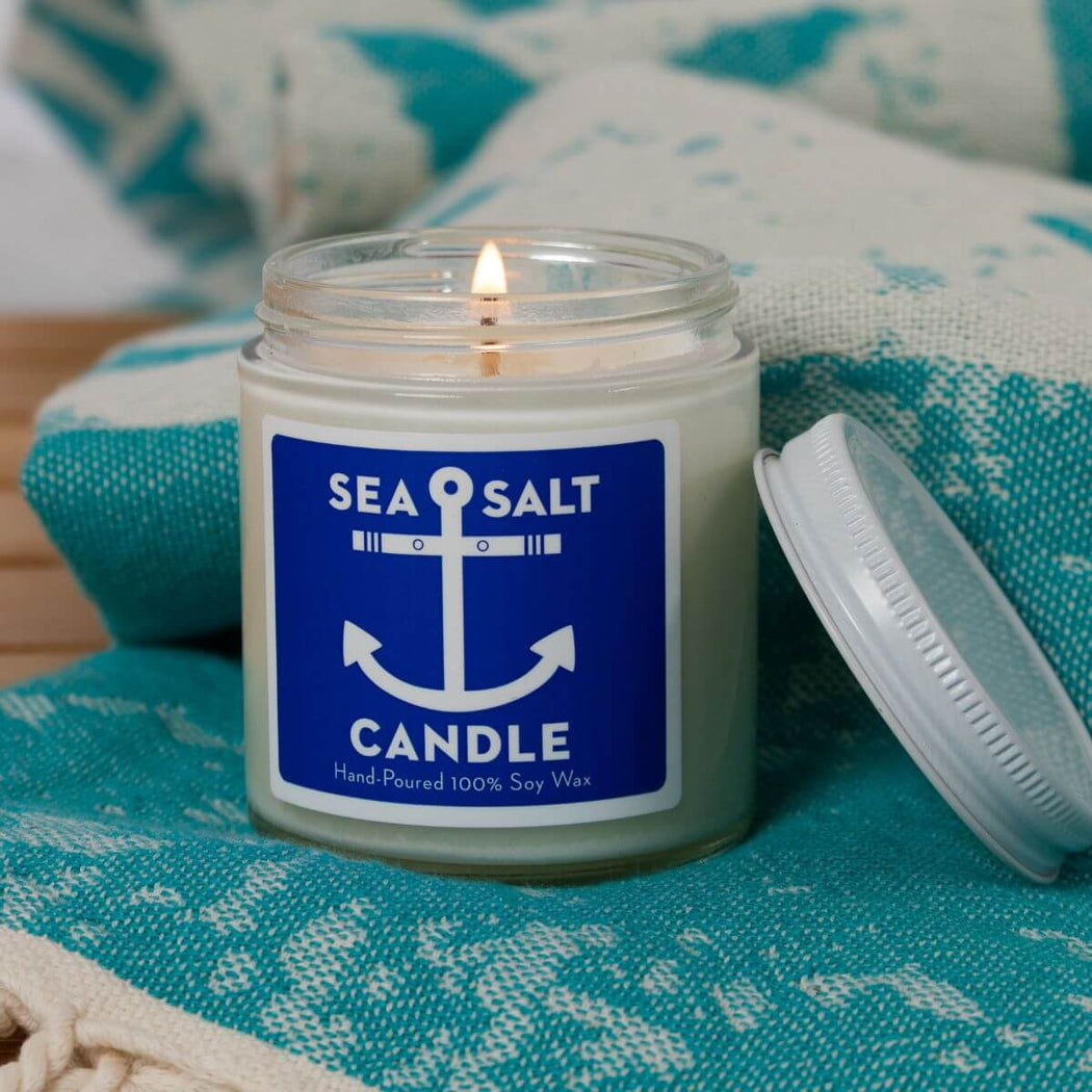 A lit sea salt candle in a clear glass jar with a blue and white anchor label, resting on a teal patterned textile with its white lid beside it.