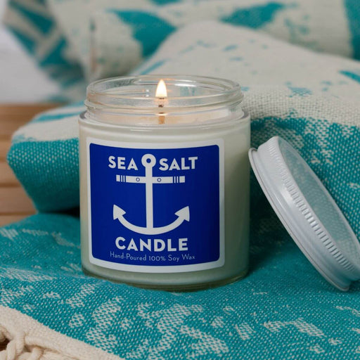 A lit sea salt candle in a clear glass jar with a blue and white anchor label, resting on a teal patterned textile with its white lid beside it.