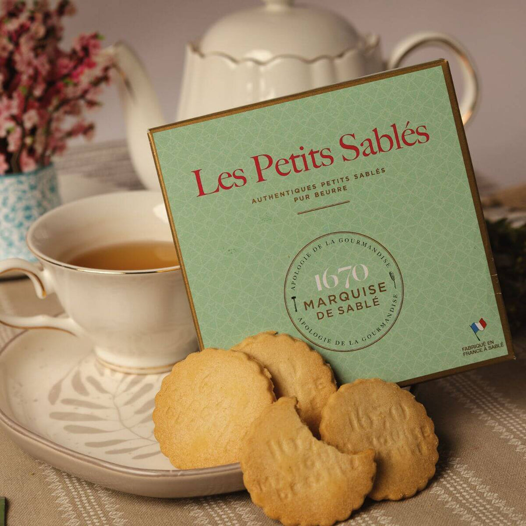 Box of 'Les Petits Sablés' cookies with a cup of tea and cookies on a table.