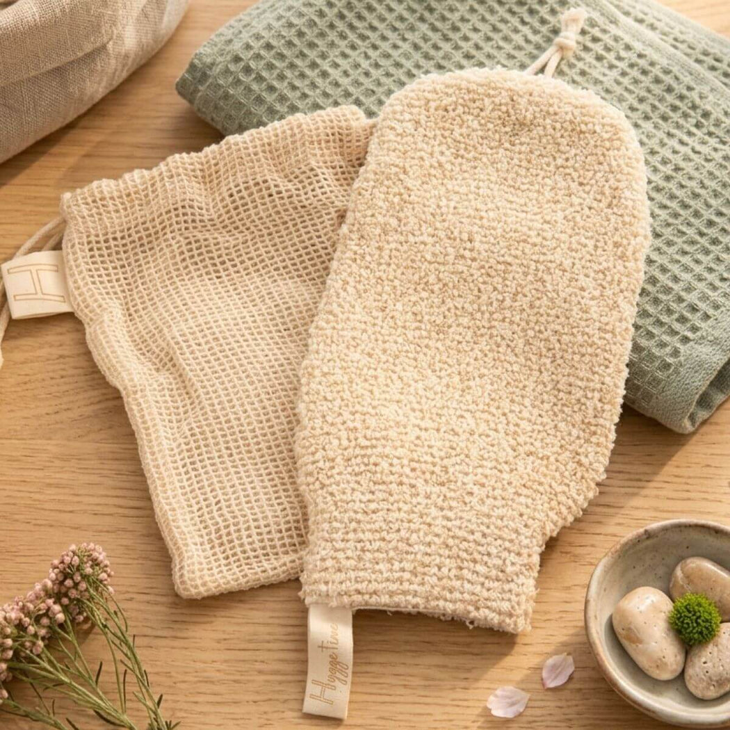 Beige textured bath mitt with a drawstring travel bag on a wooden surface with a small bowl and flowers.