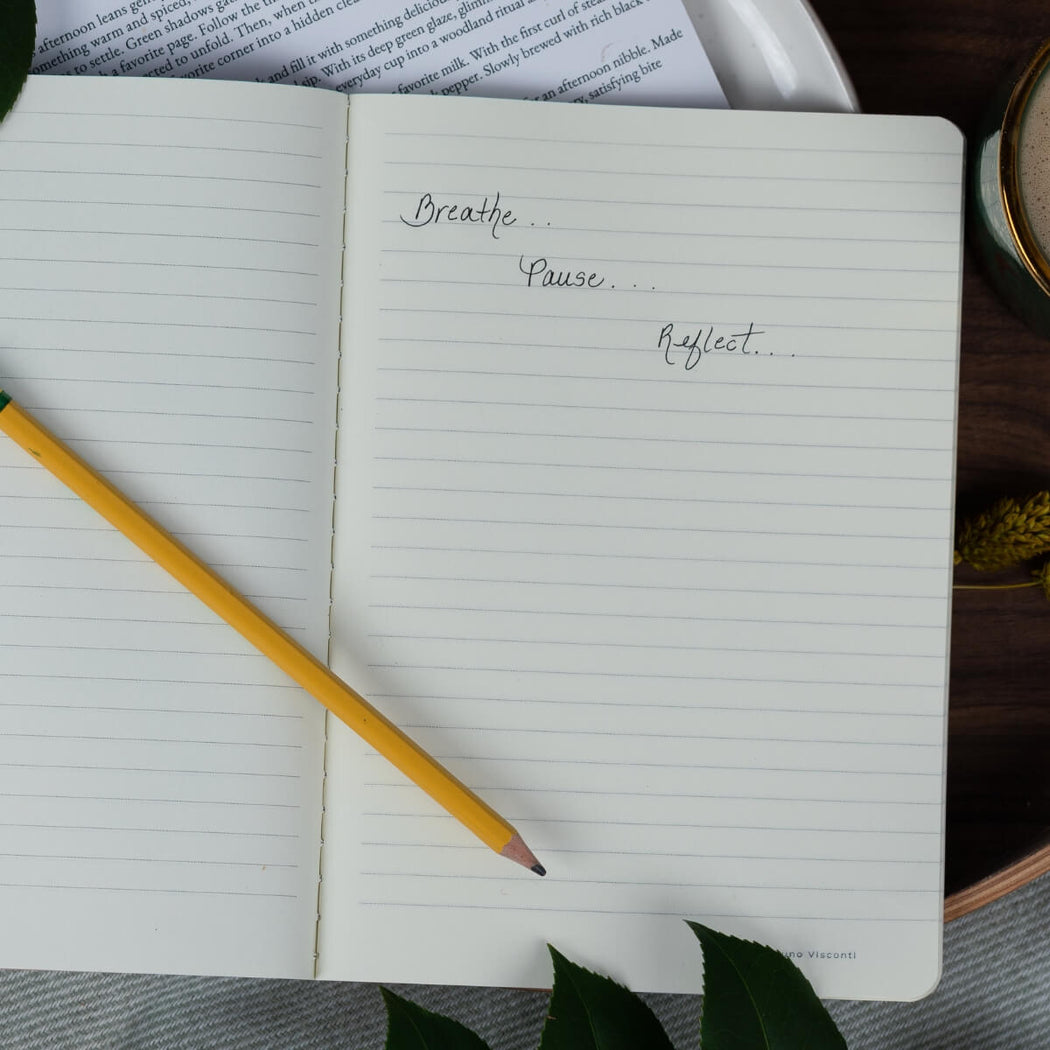 An open lined notebook with handwritten words on the right page, styled on a dark wooden surface with a yellow pencil, leaves, and a plate in the background.