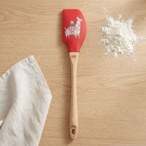 A red silicone spatula with a llama illustration and wooden handle, laid flat on a light oak surface beside a linen towel.