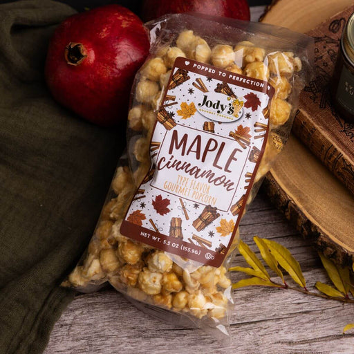 Bag of Jody's Maple Cinnamon popcorn on a wooden surface with a pomegranate and autumn leaves.