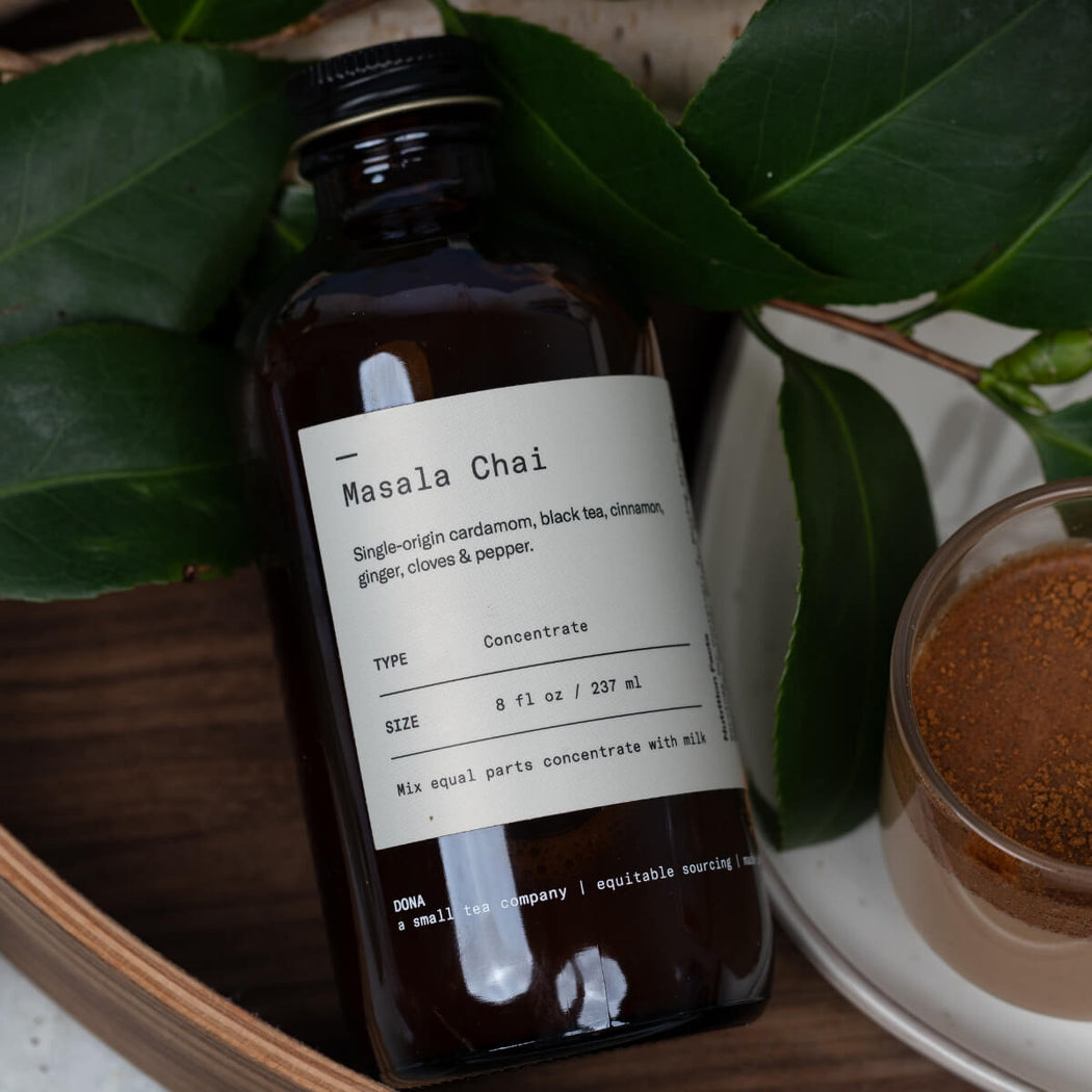 An amber glass bottle labeled 'Masala Chai' with a black cap, styled on a wooden surface with green leaves and a small glass of warm brown powder beside it.