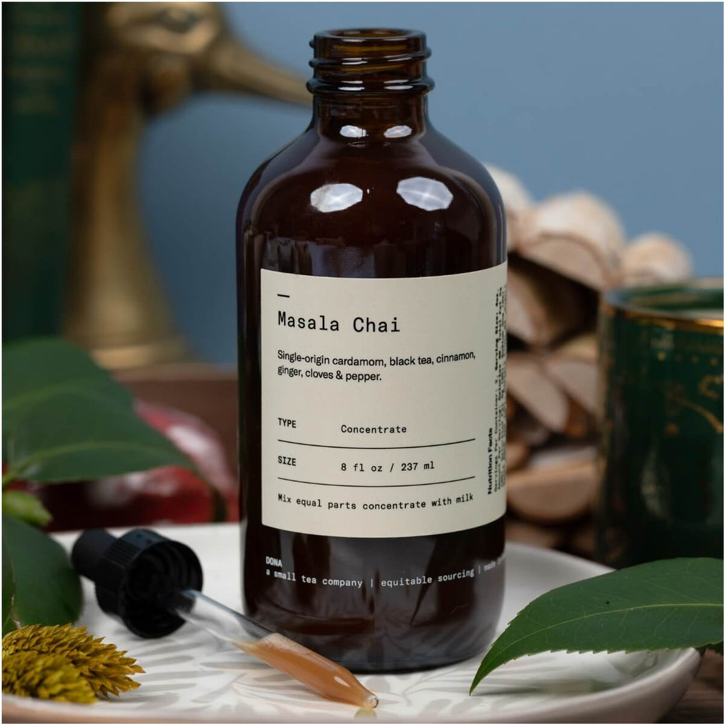 An amber glass bottle of Masala Chai tea concentrate with a white label, styled on a white ceramic dish with a glass dropper and green leaves against a soft blue background.