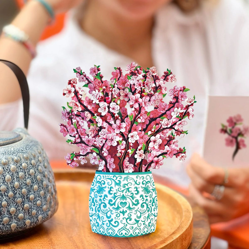 Decorative pop-up paper greeting card of a pink floral tree in a white pot with blue patterns on a wooden table.