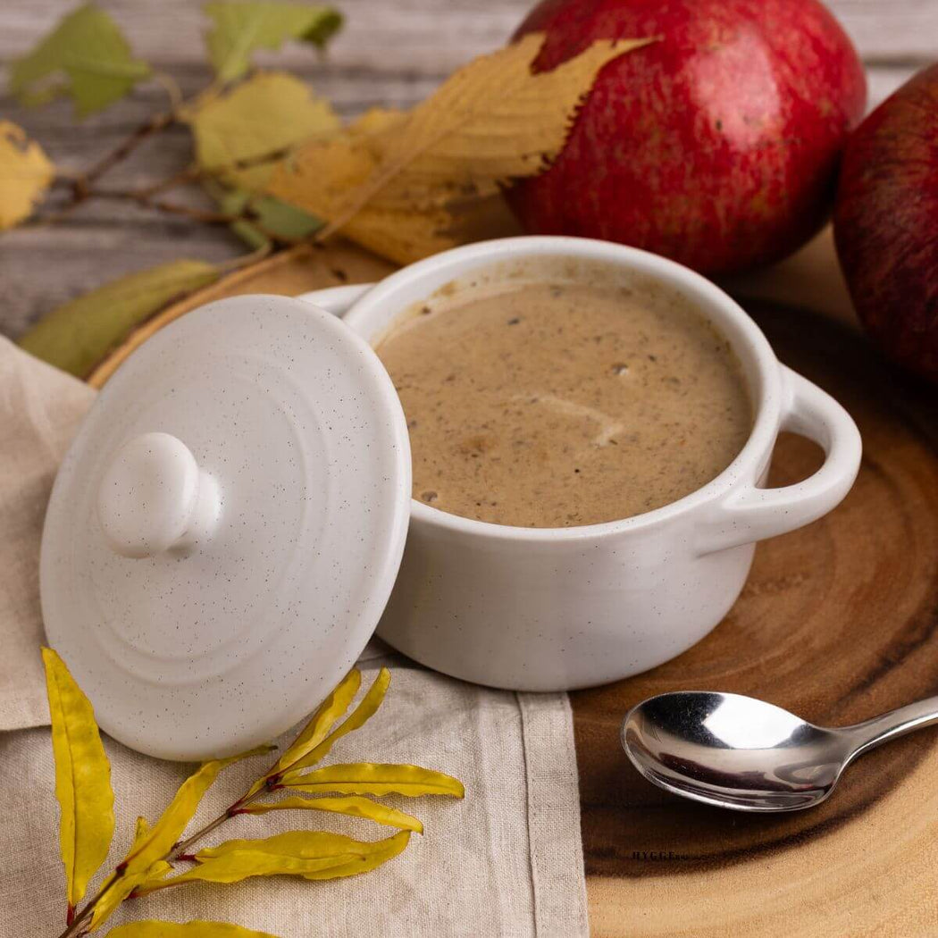 Mini round white cocotte with a lid containing creamy soup on a wooden surface with autumn leaves and apples.