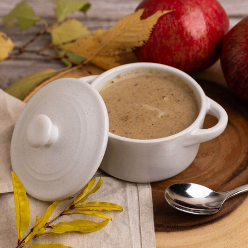 Mini round white cocotte with a lid containing creamy soup on a wooden surface with autumn leaves and apples.
