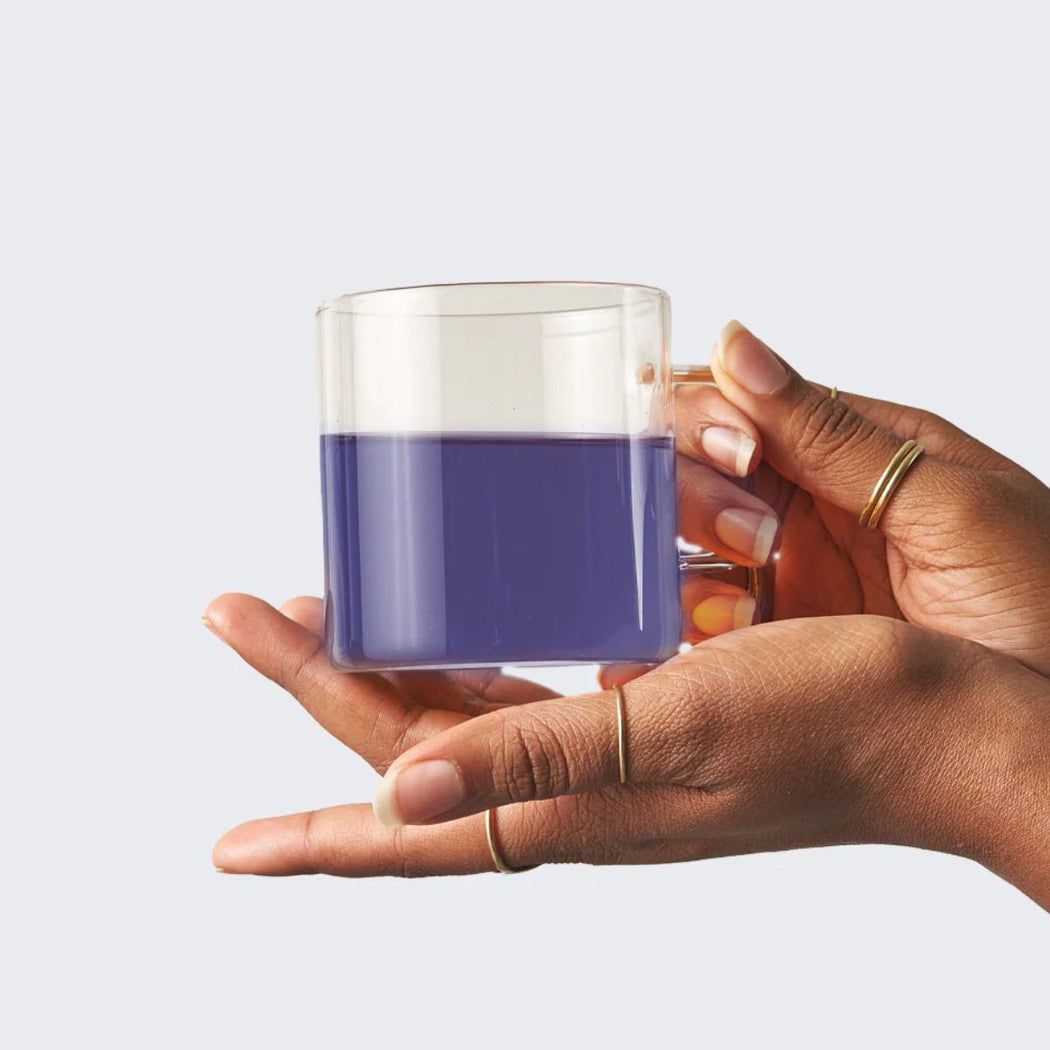 Hand holding a clear glass mug with butterfly pea tea against a white
