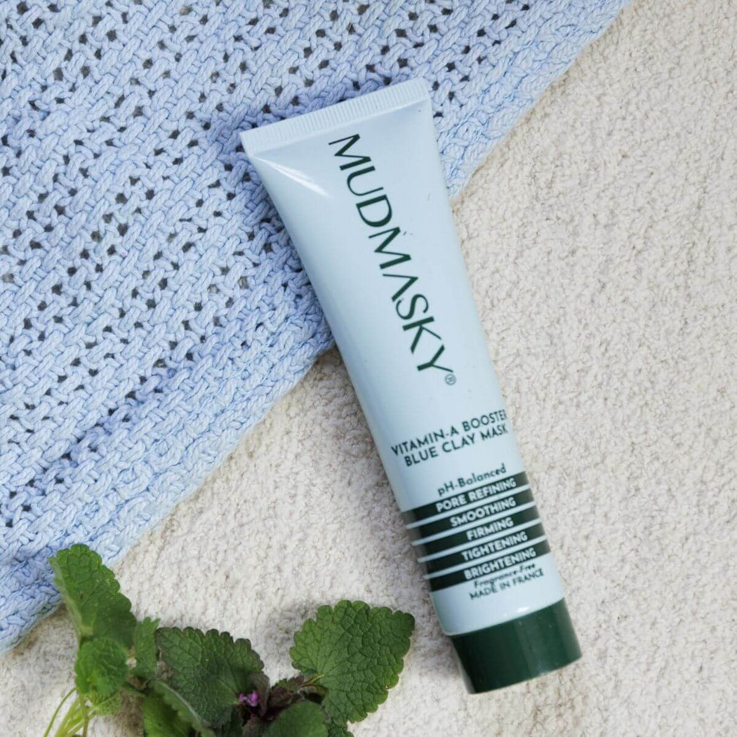 Mudmasky vitamin A booster blue clay mask tube on a textured surface with green leaves.