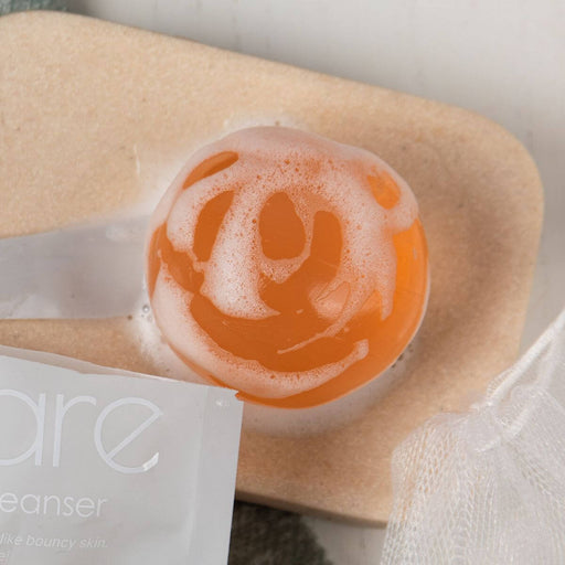 Peachy Pink Aqua Jelly Ball Cleanser lathering with foam on a soap dish beside packaging and mesh pouch.