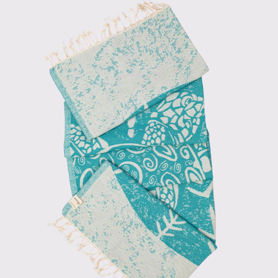 A folded turtle patterned peshtemal with ivory fringe, featuring a turquoise and off-white design with a small woven label.