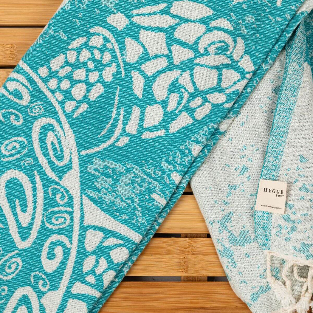 A folded turquoise and white sea turtle patterned towel with fringed edges, displayed on a light wood slatted surface. A small woven label reads 'HYGGE BOX'.