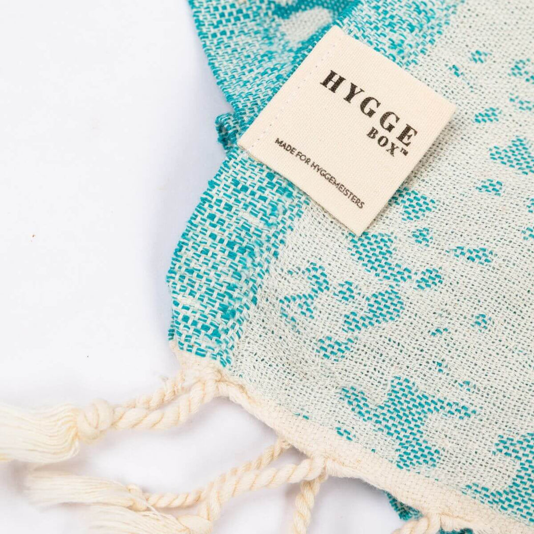 A close-up of a turkish towel with cream tassels and a cream brand label reading 'HYGGE BOX'. The towel has a turquoise and cream turtle pattern on a white background.
