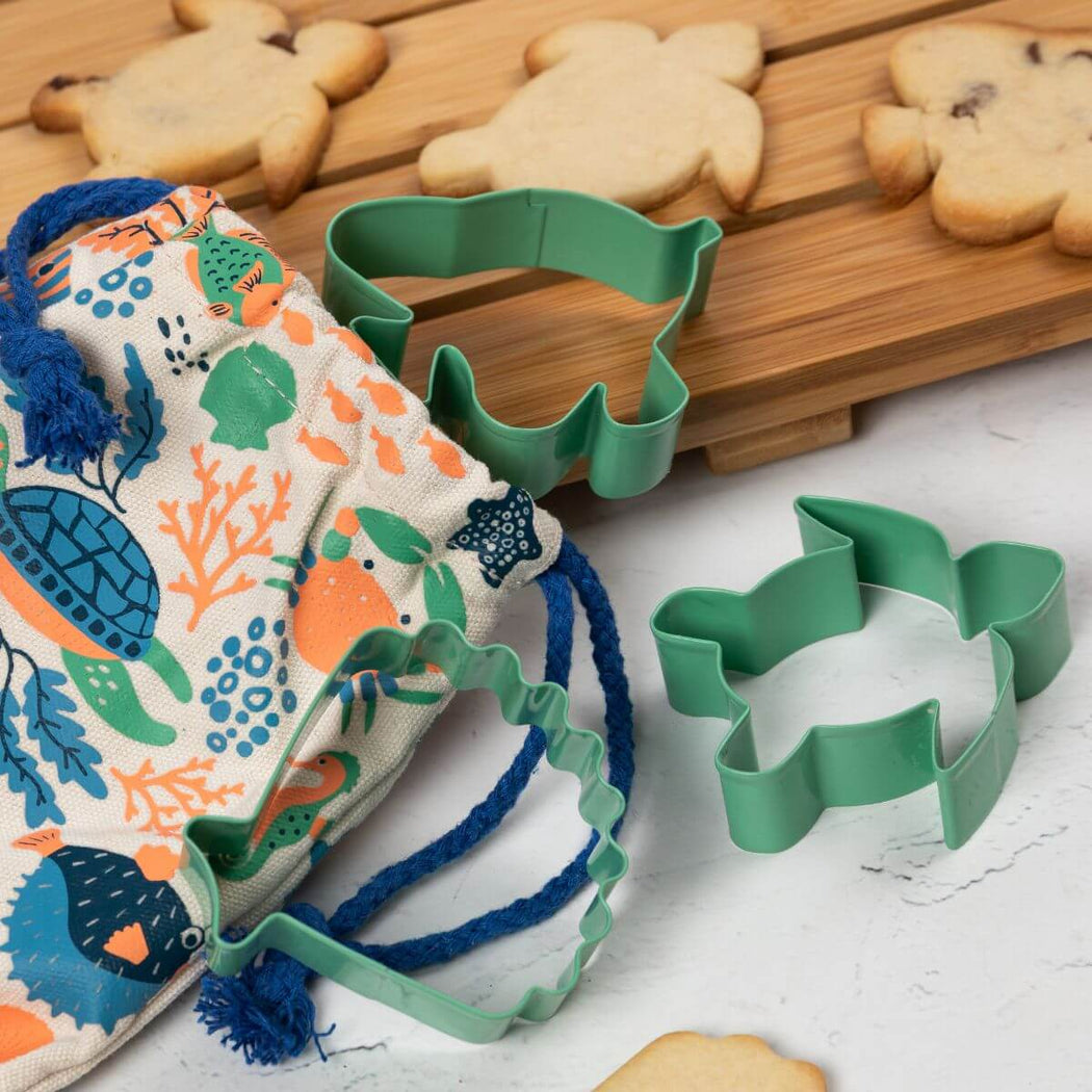 Sage green cookie cutters, including a fish-shaped cutter, shell-shaped cutter and a turtle-shaped cutter, displayed alongside a colorful fish-themed drawstring bag on a white surface with wooden boards and cookies in the background.