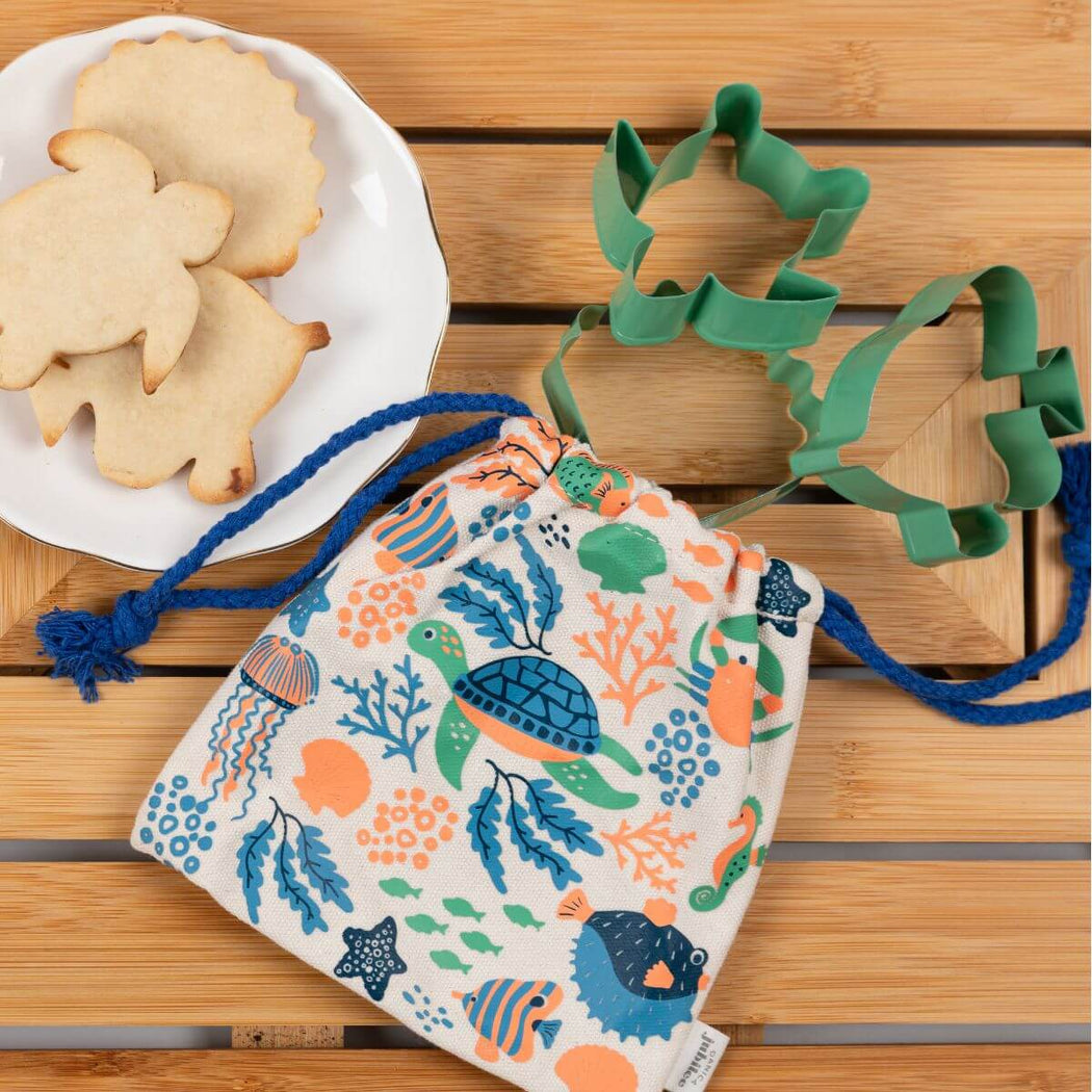 A fabric drawstring bag with an ocean-themed print of sea turtles, fish, and coral in blue, green, and orange on a white background, styled on a wooden table with sea theme cookie cutters and cookies.