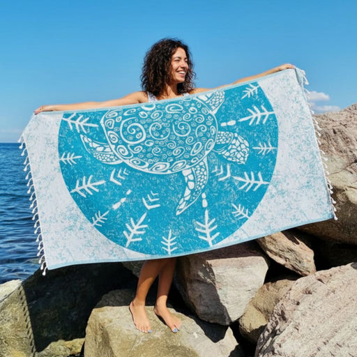 Woman standing on rocks on a clear sunny day holding a turquoise turkish towel with a sea turtle print.