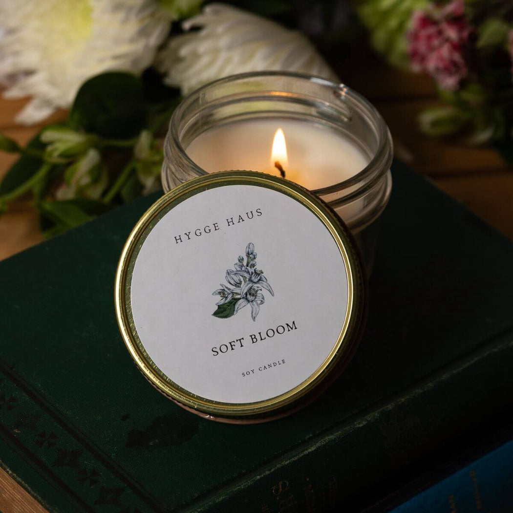 Candle in a glass jar with a visible label on a dark surface with flowers in the background