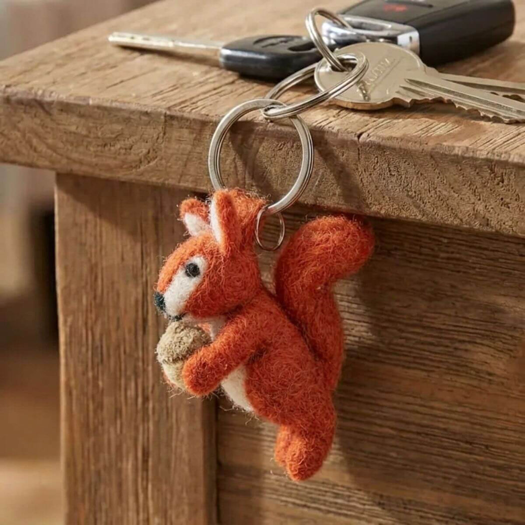 A felted orange-red squirrel keychain holding an acorn, attached to a metal keyring with keys resting on a wooden table.