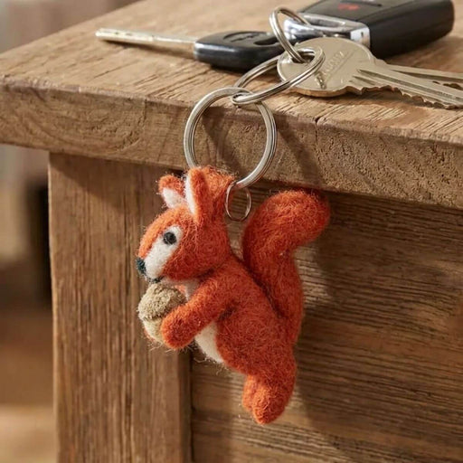 A felted orange-red squirrel keychain holding an acorn, attached to a metal keyring with keys resting on a wooden table.