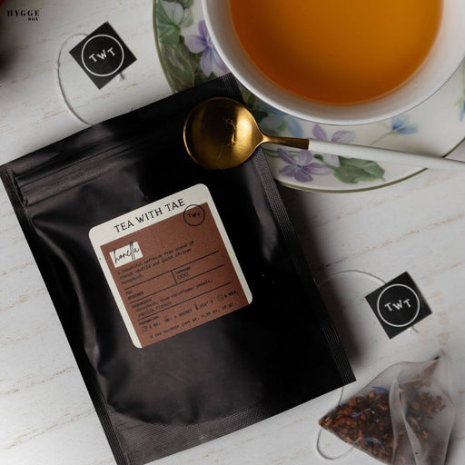 Tea packaging with a label, a cup of tea, and a spoon on a white surface.