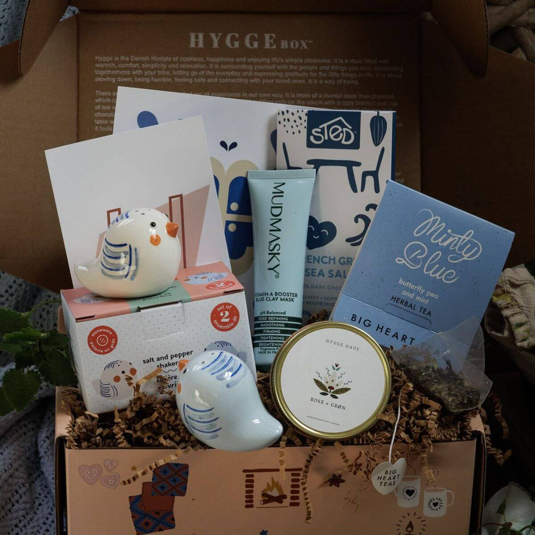Assorted items in a curated Hygge Box including a candle, chocolate bar, clay mask, tea, and birdie salt and pepper shakers on a textured surface.