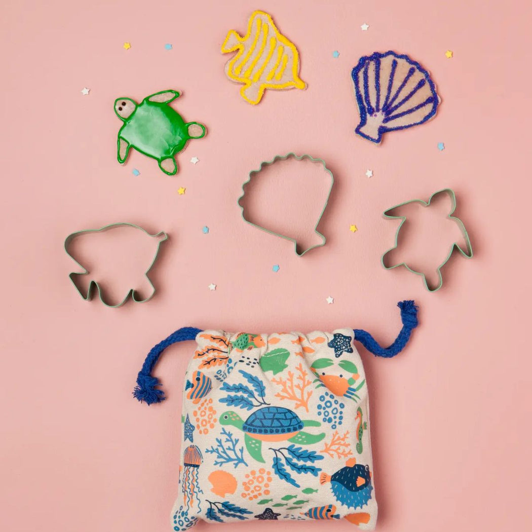 A 3 piece set of metal cookie cutters shaped like a turtle, fish, and shell, arranged on a pink background above a matching fabric drawstring pouch with an ocean-themed print.
