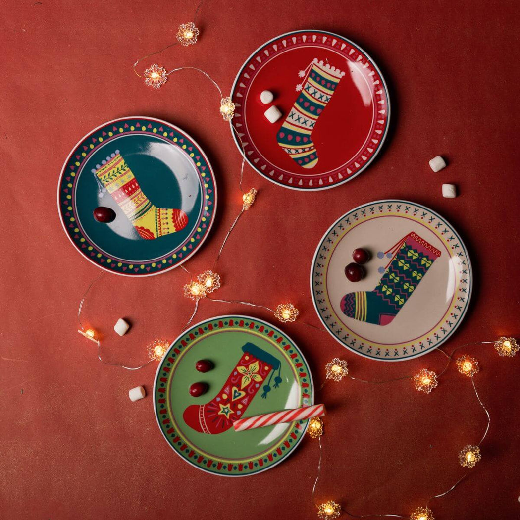 Four decorative plates with festive stocking designs on a red background with string lights.