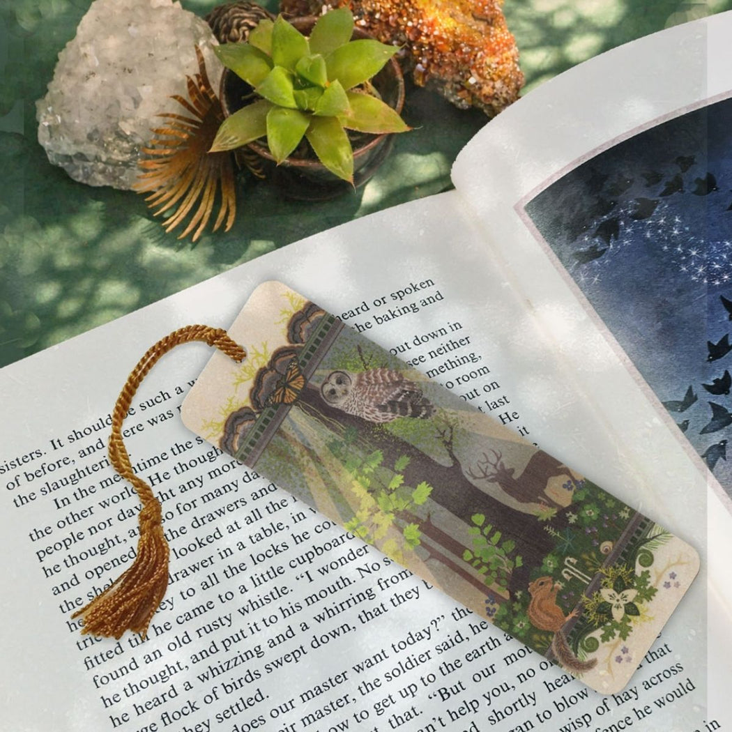 A rectangular illustrated wooden bookmark with a mustard color braided cord, placed on an open book. The wooden bookmark features a woodland scene with a owl, trees, and small illustrations.