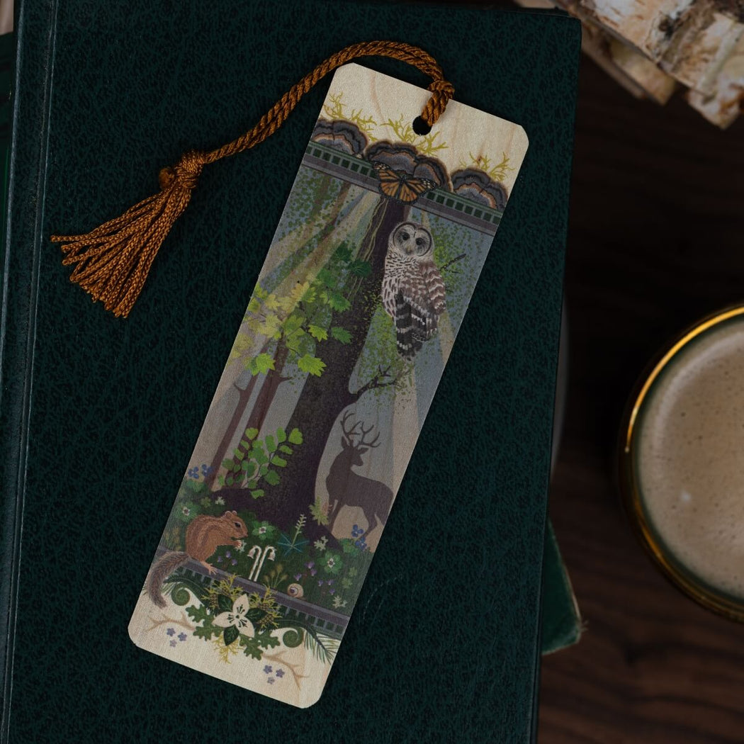 A decorative wooden bookmark with a mustard color tassel, featuring an illustrated forest scene with an owl, deer, chipmunk, and flowers, resting on a dark green textured book.