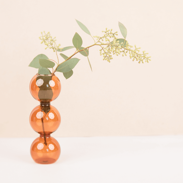 A glass bubble vase with three spherical segments in amber color, filled with water and displaying a small branch with green leaves.